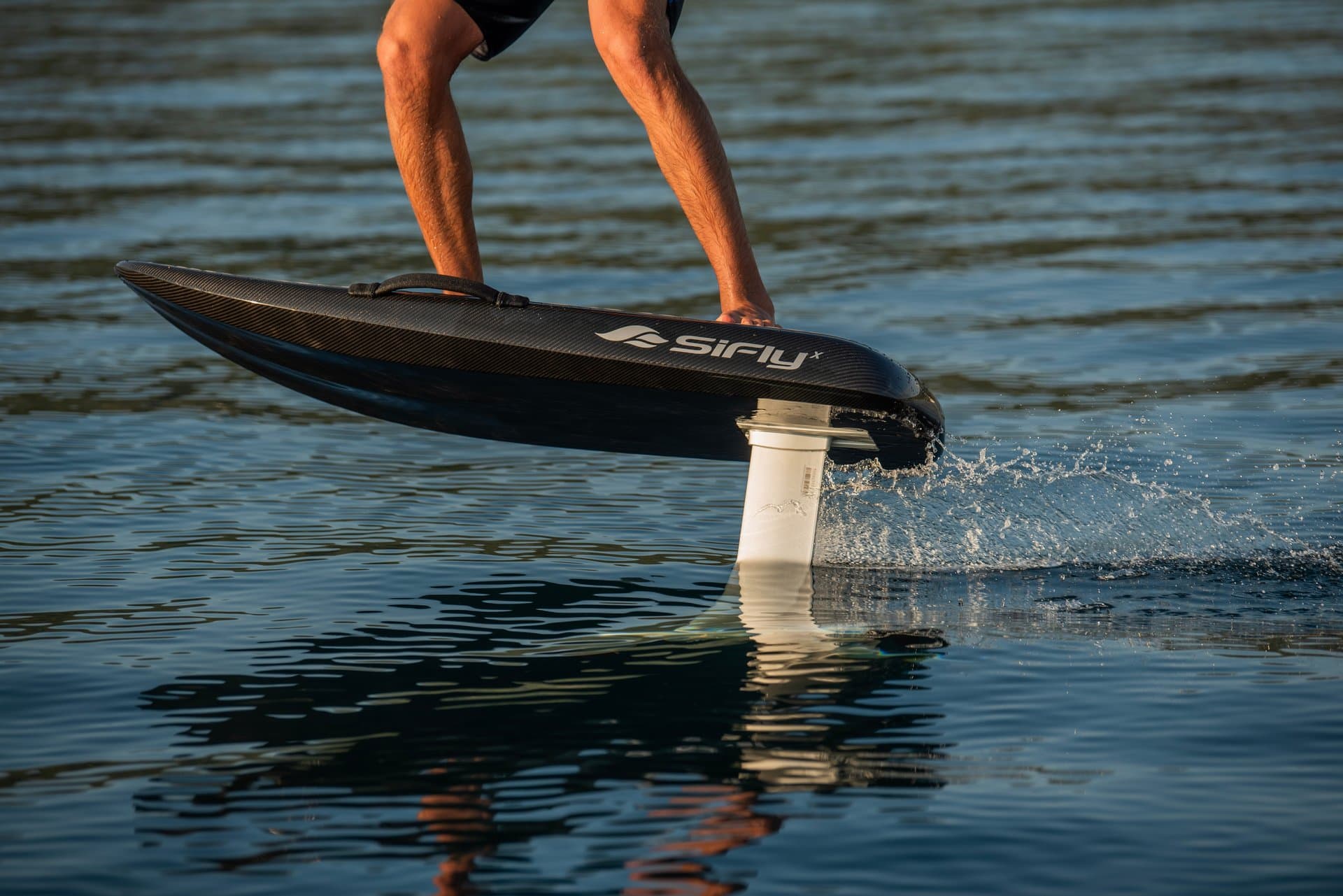 Close-up of rider's legs on carbon SiFly X eFoil board hydrofoiling above calm water, white mast cutting surface with splash