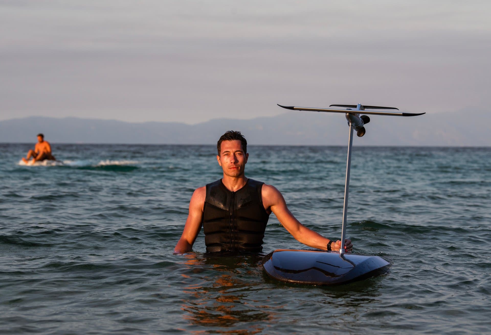 Man in black sleeveless neoprene vest standing chest-deep in ocean, holding SiFly eFoil board with mast and foil wing raised,