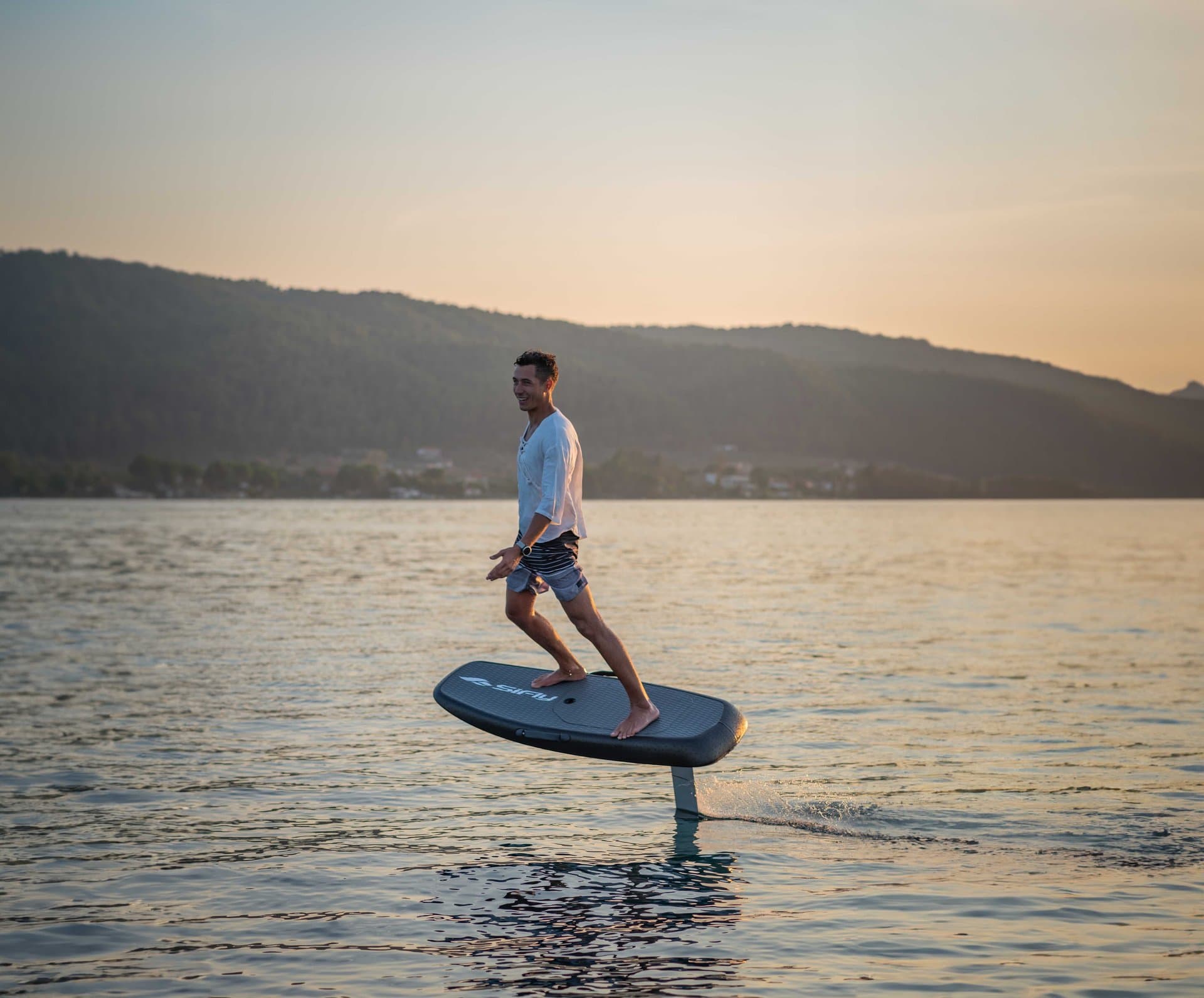 Smiling man in white shirt and shorts hydrofoiling above calm lake on a SiFly eFoil board at golden-hour sunset, hills behind