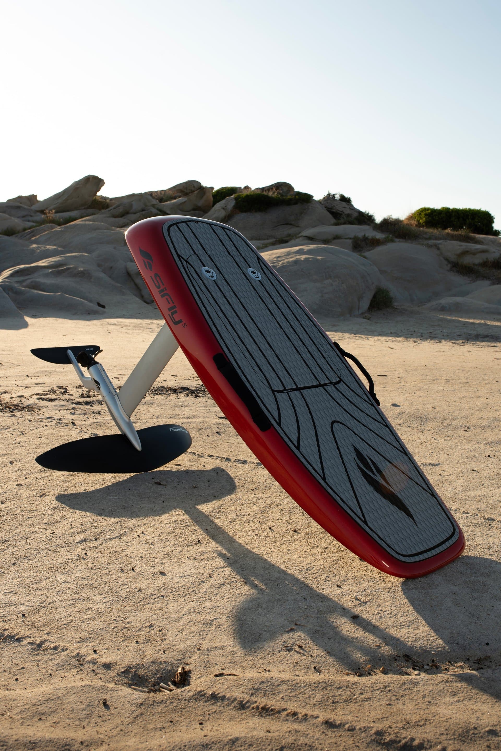 SiFly eFoil red board with grey teak deck pad and hydrofoil assembly propped upright on sandy beach, rocky coastal boulders b