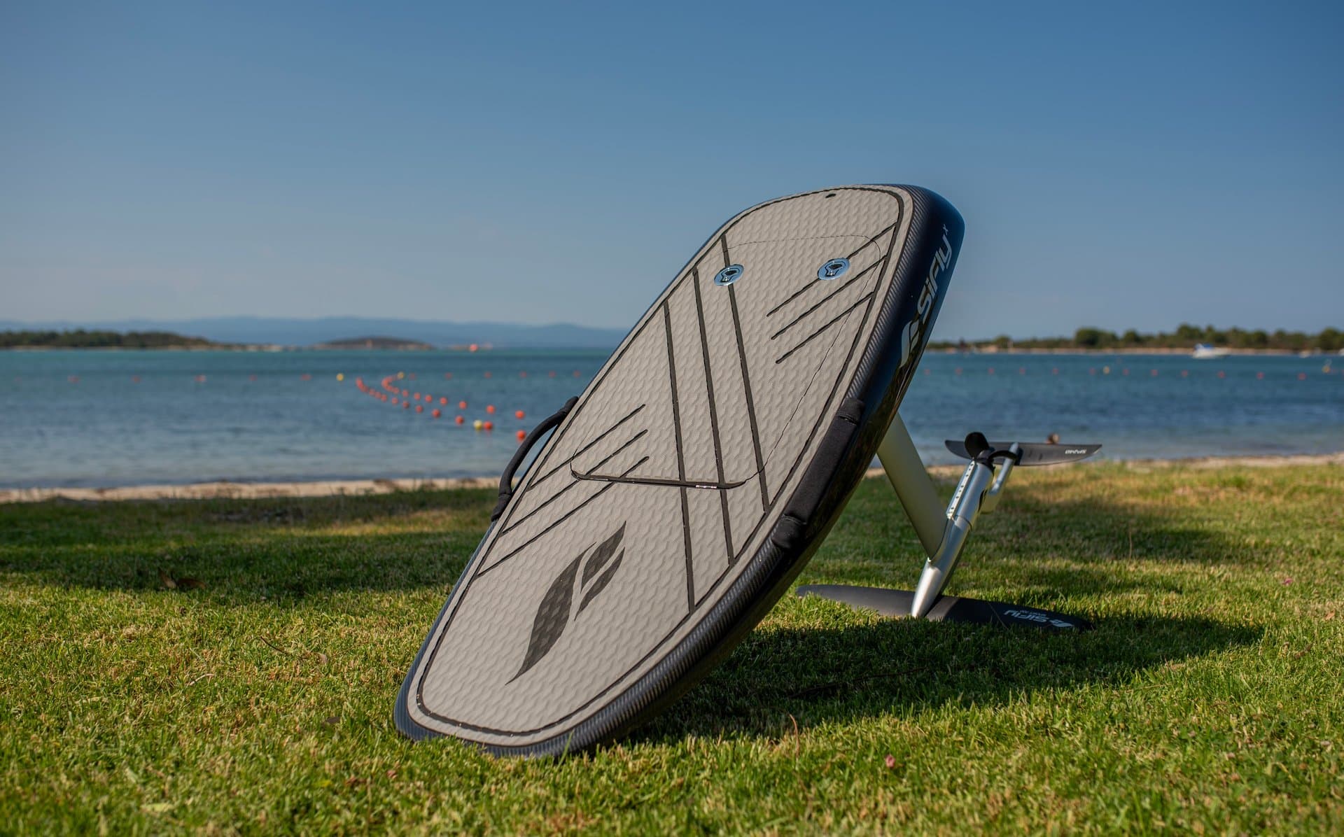 SiFly eFoil board leaning upright on grass showing textured deck pad, mast, and propeller, calm lake and buoy line behind
