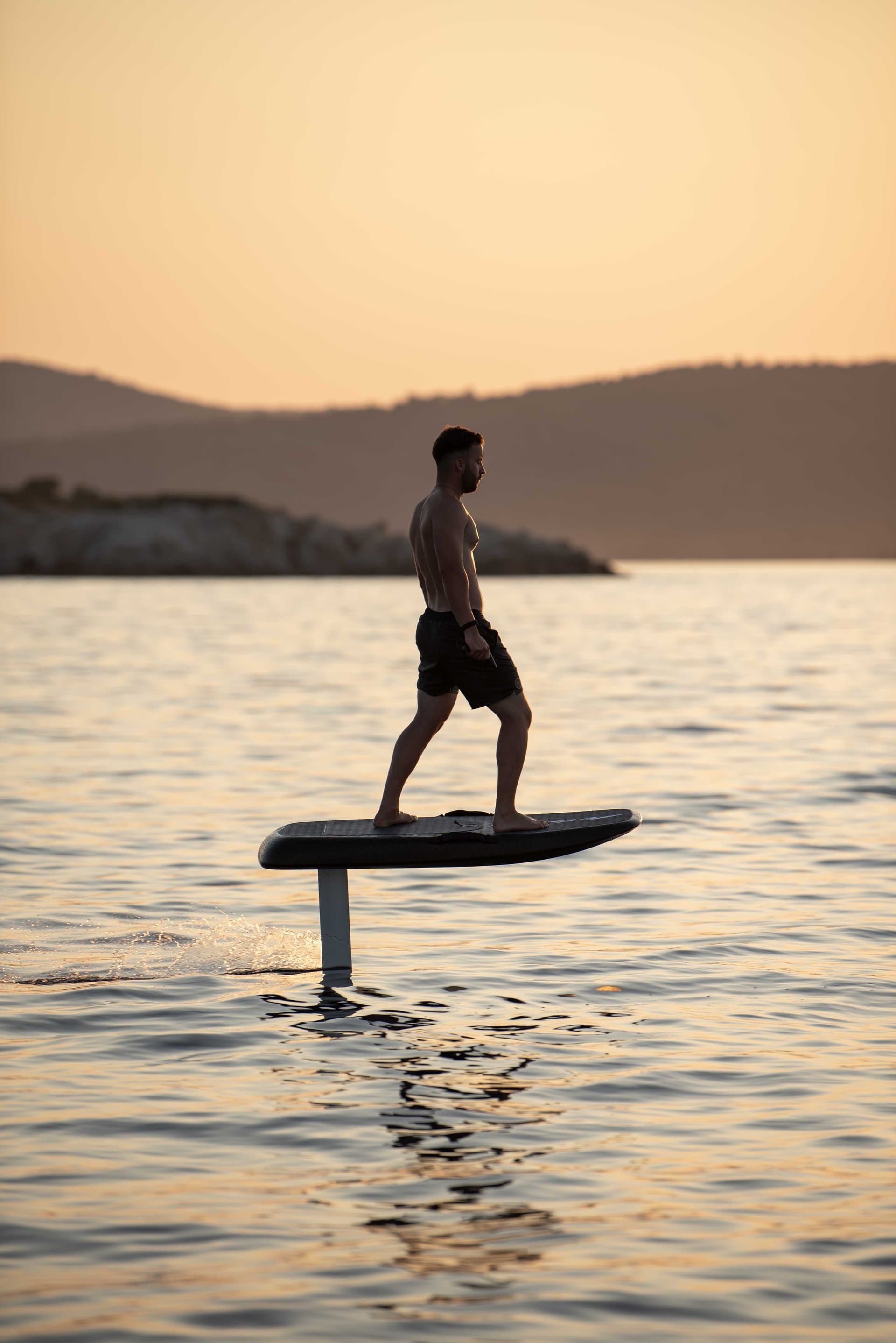 Shirtless man in black shorts riding a SiFly eFoil board, foiling above calm sea at sunset with hilly coastline silhouetted b