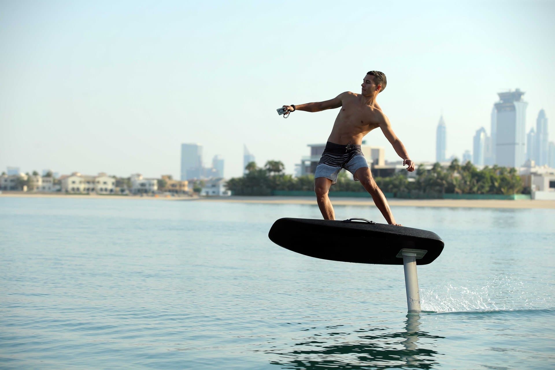Shirtless man with outstretched arm hydrofoiling on a dark SiFly eFoil board above calm bay, Dubai high-rise skyline visible 