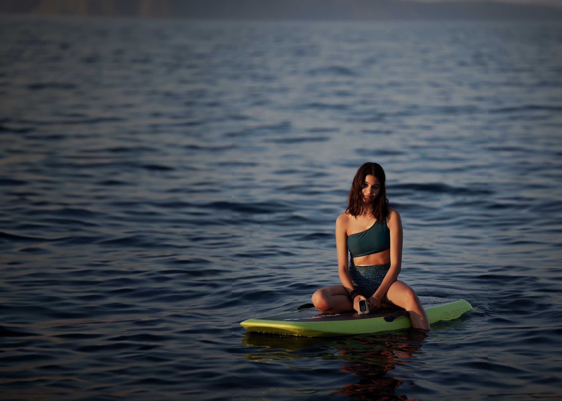 Young woman in bikini top seated cross-legged on a green SiFly eFoil board floating on calm open water at golden hour, holdin