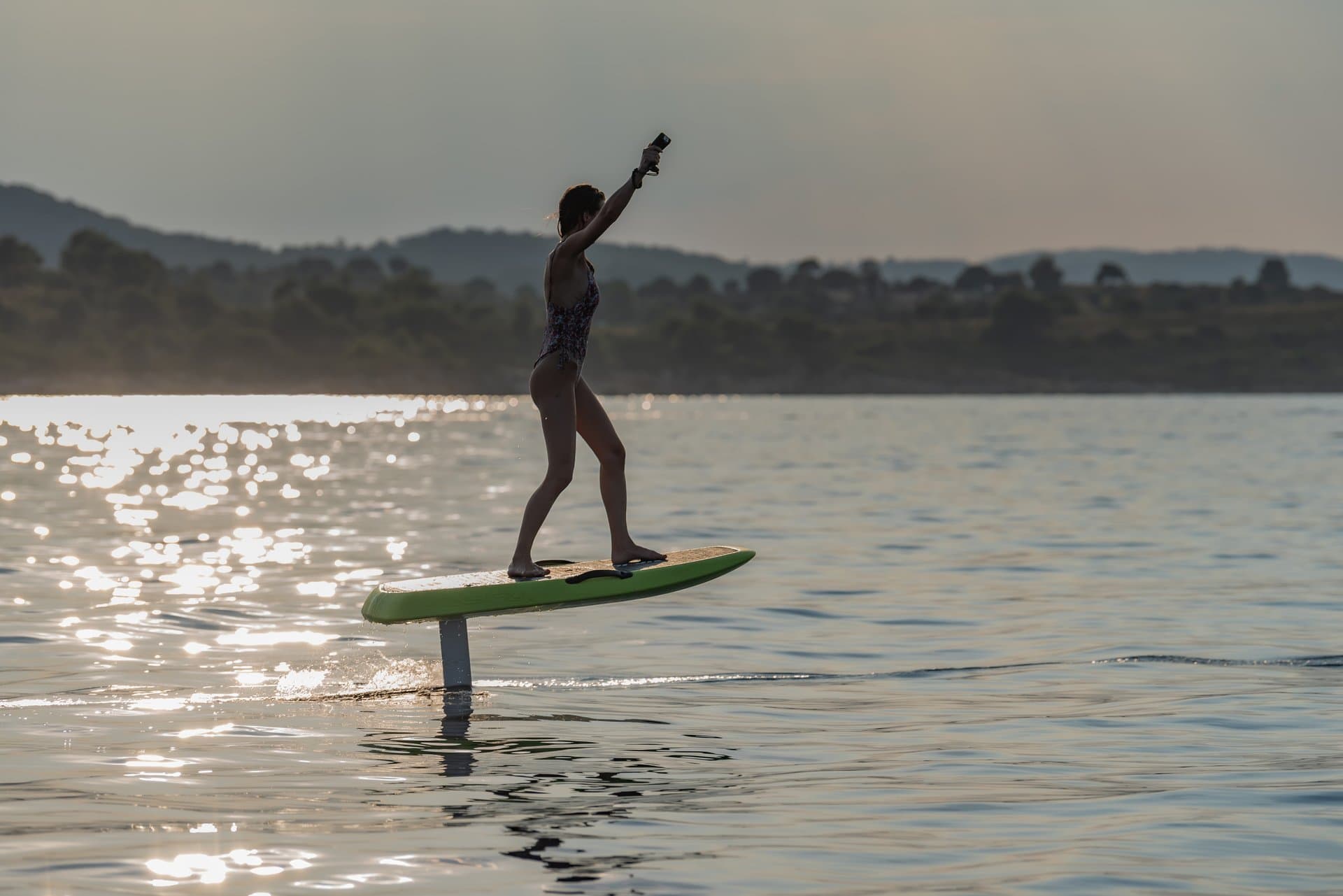 Woman in one-piece swimsuit raising arm with camera while hydrofoiling on a green SiFly eFoil board above calm lake at sunset