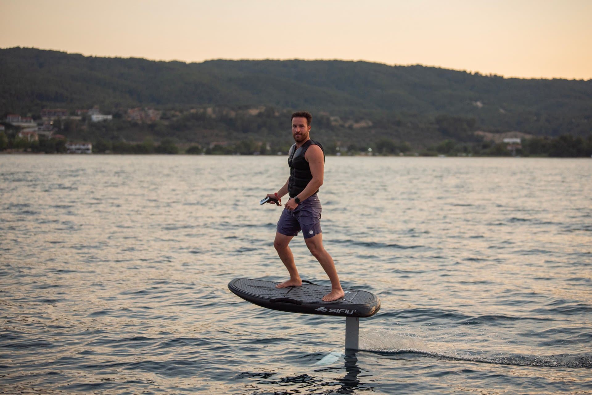 Upright man in black vest and shorts foiling on SiFly eFoil at dusk, turning to look back, forested hills and lakeside villag