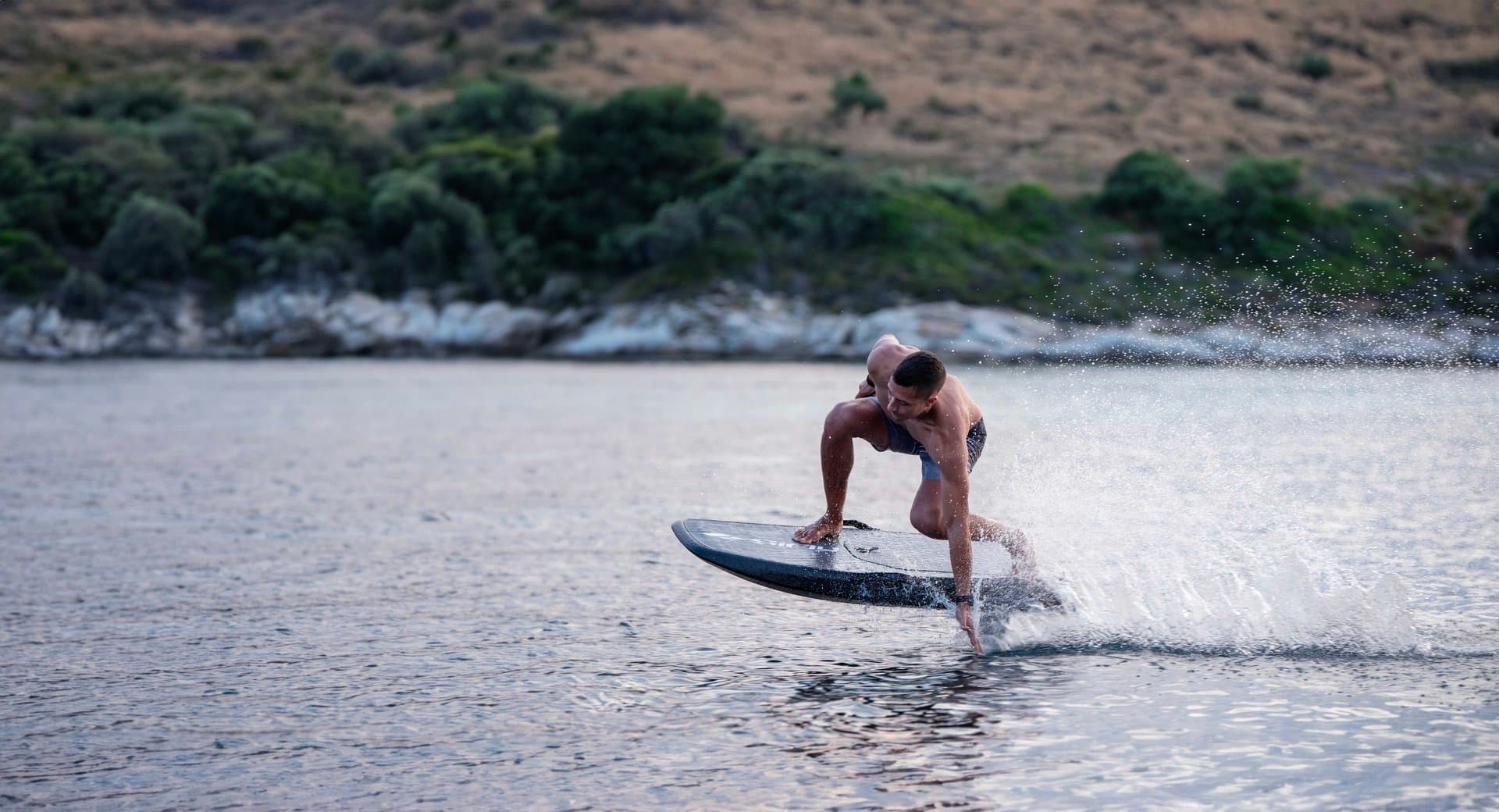 Crouching shirtless man touching SiFly eFoil board deck while skimming calm bay at speed, dramatic water spray, tree-lined ro