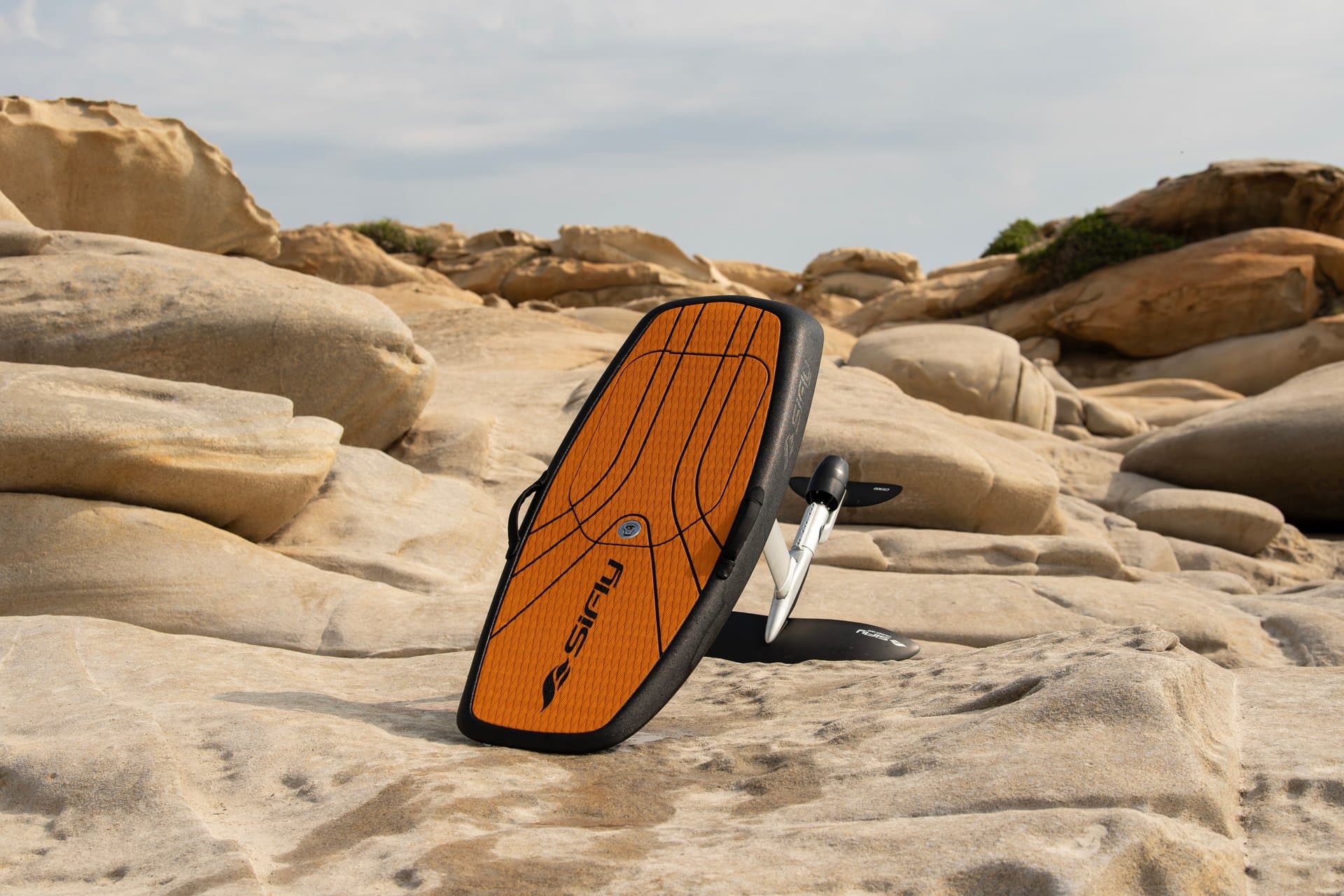 SiFly eFoil board with orange grip deck pad and white hydrofoil mast standing on sandy coastal rock shelf, dramatic low-angle