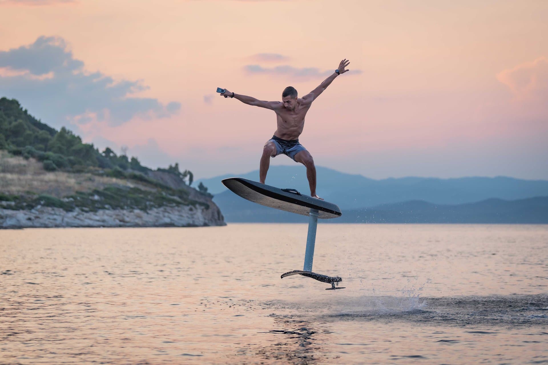 Shirtless man arms outstretched balancing on a SiFly eFoil, hydrofoiling above calm coastal sea at dusk, rocky cliffs and mou